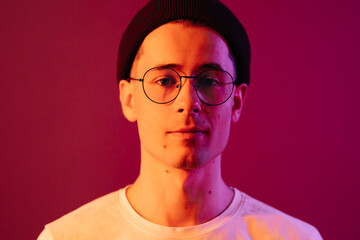 Shot of young handsome male with glasses trendy hairdo, wears casual white t shirt, has positive expression, poses in studio against pink background splashed by colorful lights