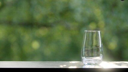 Empty clean glass on the brown organic wooden table with green morning summer or spring leaves of trees bokeh background. Sun's rays glare on a glass beaker
