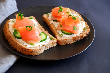 Toast with red fish, curd cheese and cucumber on a black plate.