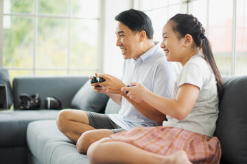 Young Asian love family father and daughter play a game consoles wireless controller sitting on sofa in living room at home which smiling and felling happy. live at home concept in COVID-19 situation