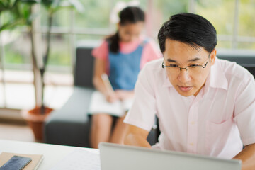 Obraz premium Young Asian love family father working use computer notebook laptop and daughter do homework on sofa in living room at home which smiling and felling happy. live at home concept in COVID-19 situation