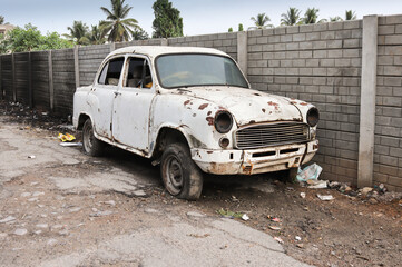 An Old abandoned car of  Indian make in dilapidated condition with lots of rust and missing parts seen on an empty street.