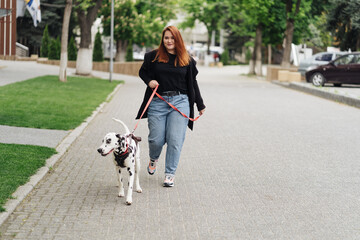 Naklejka na ściany i meble View of young Caucasian female cynologist walking at city urbanity during morning time for training dalmatian dog, plus size female spending leisure with pet best friend enjoying weekend at street