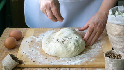 The hands of an elderly woman knead the dough for bread on a wooden board on the table, on the table there are 2 eggs, there are linen bags with spices cumin and coriander and flour. World Bread Day