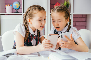 Two young female classmates are sitting at a desk and using smartphones. Modern technology concept.