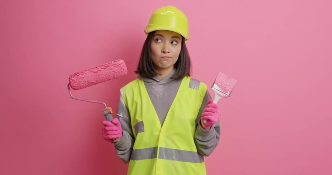 Home improvement. Serious thoughtful Asian female decorator or builder holds roller and brush going to paint walls wears protective helmet and uniform poses with painting tools against pink background