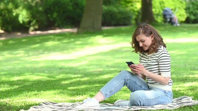Pretty young woman relaxing in park with cellphone and chatting with friend at summer day