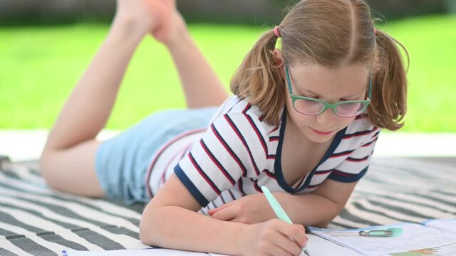 Pretty teen girl in glasses with pigtails laying on blanket in backyard with green grass and study