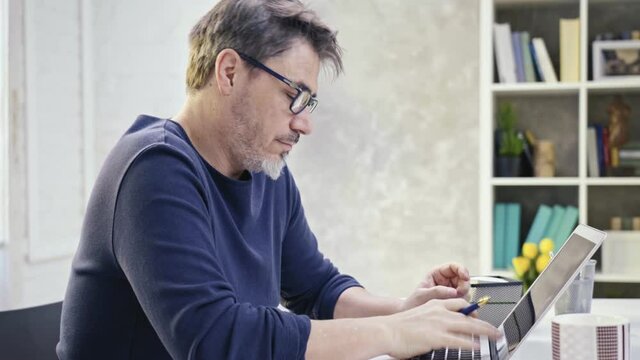 Man sitting at desk at home with laptop computer. Businessman working in home office. Portrait of mature age, middle age, bearded, glasses, authentic look.