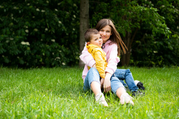 Young woman mom is sitting on green grass in a summer park with a boy son. Mom hugs her boy son.