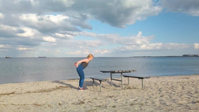 Young caucasian woman doing gym exercises and training on the beach in Amager, Copenhagen, Denmark. The girl is doing box jumps on a bench. It is a sunny spring evening. 3x times speeded up 4K clip 