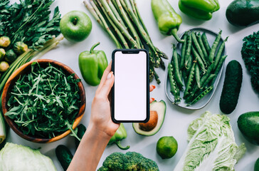 Woman hand holding smartphone with white screen mock-up, in front fresh vegetables on the table....