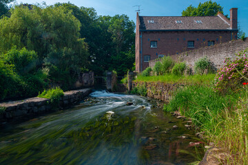 Fototapeta na wymiar Long exposure of a water rapid of the Nekummer watermill in the Jeker river in the valley with blooming wild flowers and a old cottage just outside Maastricht