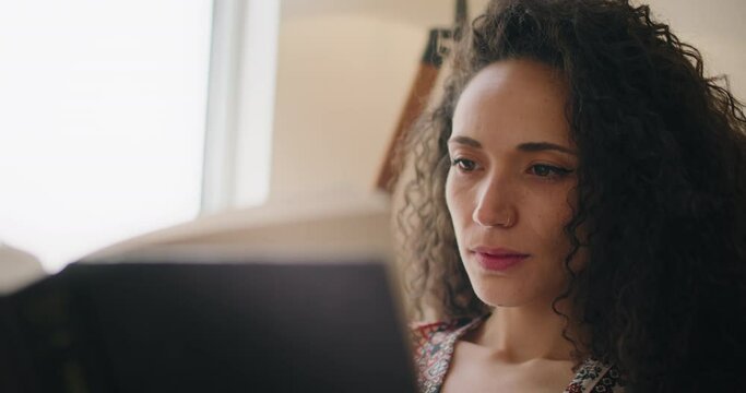 Close up of a young woman with curly hair reading a book at home. Slow motion, handheld. 