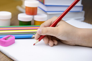 The child holds a pencil in his hand. The child is doing homework. Books, paints,pencils and a sharpener on the table