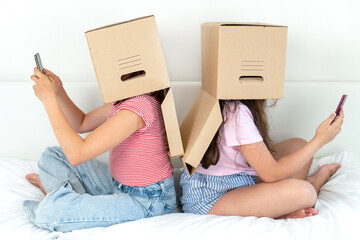 Two teenage girls with boxes on their heads are sitting and looking at smartphones on the bed at home. The concept of children's dependence on phones, social networks and games on the phone