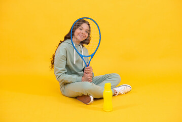 happy child sit in sportswear with squash racket and water bottle on yellow background, childhood