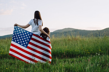 Back view of loving young mom and cute little preschooler daughter with american flag in wind...