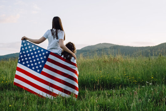 Back View Of Loving Young Mom And Cute Little Preschooler Daughter With American Flag In Wind Together Standing On Green Field On Background Summer Mountains, Enjoying Sunset On Nature. July Fourth