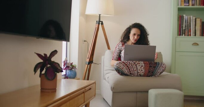 A young woman sitting on a couch with a glass of wine in hand, working on a laptop. Slow motion, handheld, medium shot. 