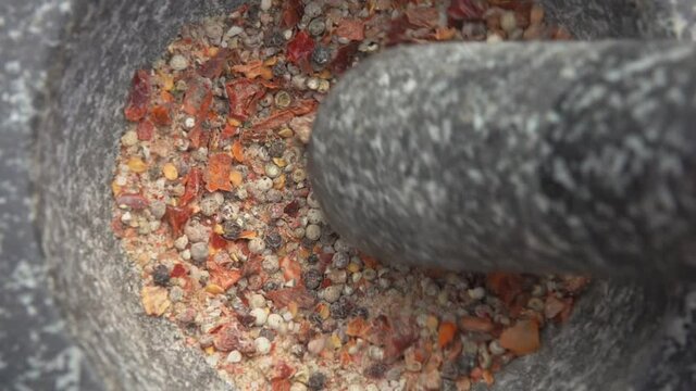 Pestle is grinding various kinds of spices and peppers in a grey stone mortar