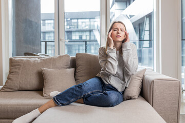 A woman holding her head anxious about an unbearable headache, sitting at home on the couch