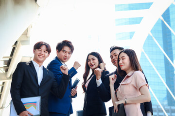 Obraz premium Group of five young Asian cheerful businesspeople raising fist hands up for celebrating success, businessman and woman cheer up their colleagues as commitment of strong teamwork at outside with skyscr