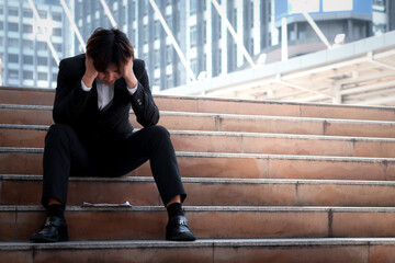 Stressed frustrated young Asian businessman sitting on stairway outside office in downtown city. man put his head in his hand thinking about his job, feeling disappointed or tired.