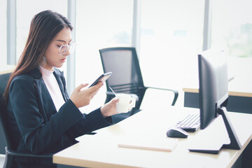 Smart and beautiful young businesswoman with black long hair in suit using mobile phone, sitting at the office desk, employee staff working at workplace.