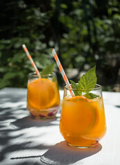 On a white wooden table in the garden, a cold orange cocktail with raspberry leaves and ice. Background - foliage from trees in the garden. Sunlight, shadow from the foliage of trees on the table.