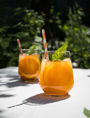 On a white wooden table in the garden, a cold orange cocktail with raspberry leaves and ice.  Background - foliage from trees in the garden. Sunlight, shadow from the foliage of trees on the table.