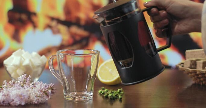 On the table, against the background of the fireplace, there is an empty cup and tea is poured into it from the teapot. Next to it is half a lemon, sugar in a cup, waffles in a cedar plate.