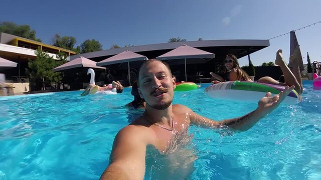 Lifestyle blogger man taking selfie video with action camera in a swimming pool. Travel vlogger films vlog from party at luxury resort. Fitted guy live streaming for social media likes. Slow motion.