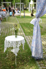In the park on the leisure area, a table covered with a tablecloth, with a bouquet of flowers, a curtain next to it, people in the distance. Background bokeh.