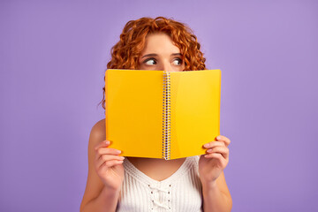 Cute redhead curly girl student teenager peeking out from behind a notebook isolated on purple background.