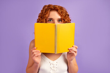 Cute redhead curly girl student teenager peeking out from behind a notebook isolated on purple background.