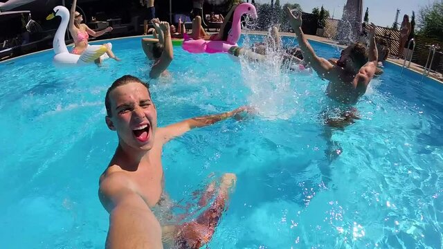 Lifestyle blogger man taking selfie video with action camera in swimming pool. Travel vlogger films vlog from party at luxury resort splashing water, having fun. Live streaming. Slow motion.