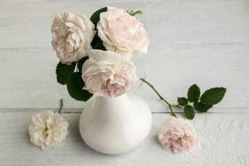 Delicate pink roses are in a white vase. The bouquet consists of three flowers. The vase is on a wooden table.