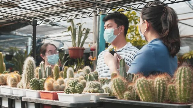 Three asian people with masks on talk about the plants around them in a nursery. Cacti plants are in the foreground. The lighting is bright.