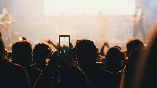 People filming rock concert on smartphones. Silhouettes crowd of people dancing at an open-air music festival. Fans applaud, raise hands up, and photographing. Party People in Action. 4K, Slow Motion