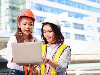 Two young beautiful Asian female engineer workers wearing safety vest and helmet, using laptop computer together, pretty women discussing about project work while walking on the way outside office in 