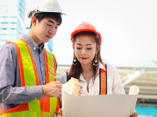 Two young Asian male and female engineer workers wearing safety vest and helmet, looking at construction blueprint together, discussing about project work while walking on the way outside office in do