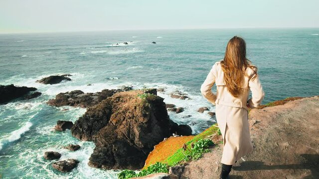 Elegantly dressed young woman in a coat walking by rocky cliffs enjoying views. Concept of a girl having a good time on vacation by exploring beautiful scenic landscapes in Cornwall, England, UK.