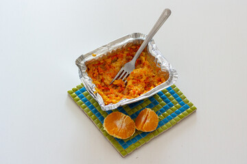 Fototapeta premium Carrot rice porridge in foil packaging and half of tangerine on a heat trivet.