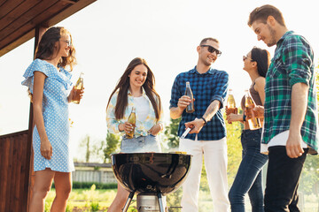 Group of people standing around a barbecue, grilling outdoors, chatting and drinking drinks, outdoors in the summer in