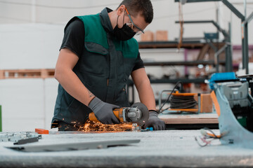 Heavy Industry Engineering Factory Interior with Industrial Worker Using Angle Grinder and Cutting a Metal Tube.He Wears a Mask on His Face Because of the Coronavirus Pandemic