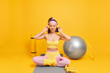 Fitness sport training concept. Sporty fit Asian woman sits crossed legs on mat enjoys listening music via headphones uses dumbbells fitball hula hoop has regular workout at home takes break