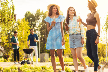 Three beautiful girlfriends having fun on a picnic, drinking drinks and chatting, a group of young friends doing barbecue on the grill