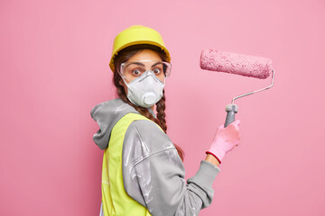 Surprised female painter moves in new dwelling busy doing repairing holds painting roller wears protective helmet respirator workwear isolated over pink background. House improvement concept