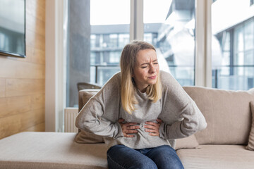 Young woman sitting on sofa holding hands on stomach twisted from pain, poisoned or menstrual pain, woman at home